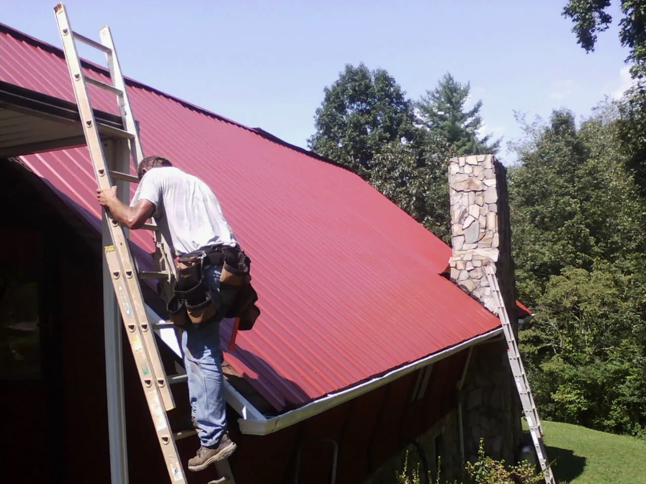Roofer on a ladder beside a red metal roof and stone chimney — Soffit & Fascia Repair by Roofing Brothers in Rockbridge